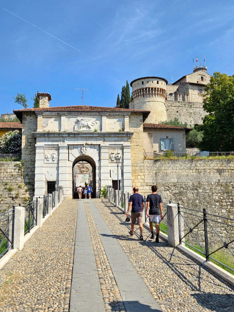 The stone entrance gate of Brescia Castle with a Venetian lion relief and visitors walking across the bridge.