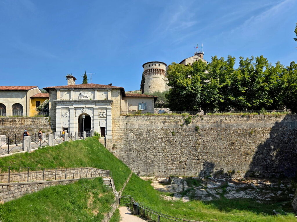 A wide view of the massive stone fortifications and entrance gate of Brescia Castle under a clear blue sky.