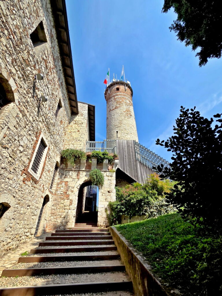 Stone steps leading up to an arched entrance and a tall medieval round tower at Brescia Castle.