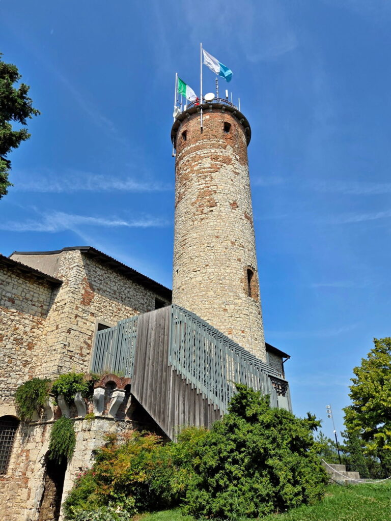 Low angle view of the Mirabella Tower at Brescia Castle with flags flying against a blue sky.