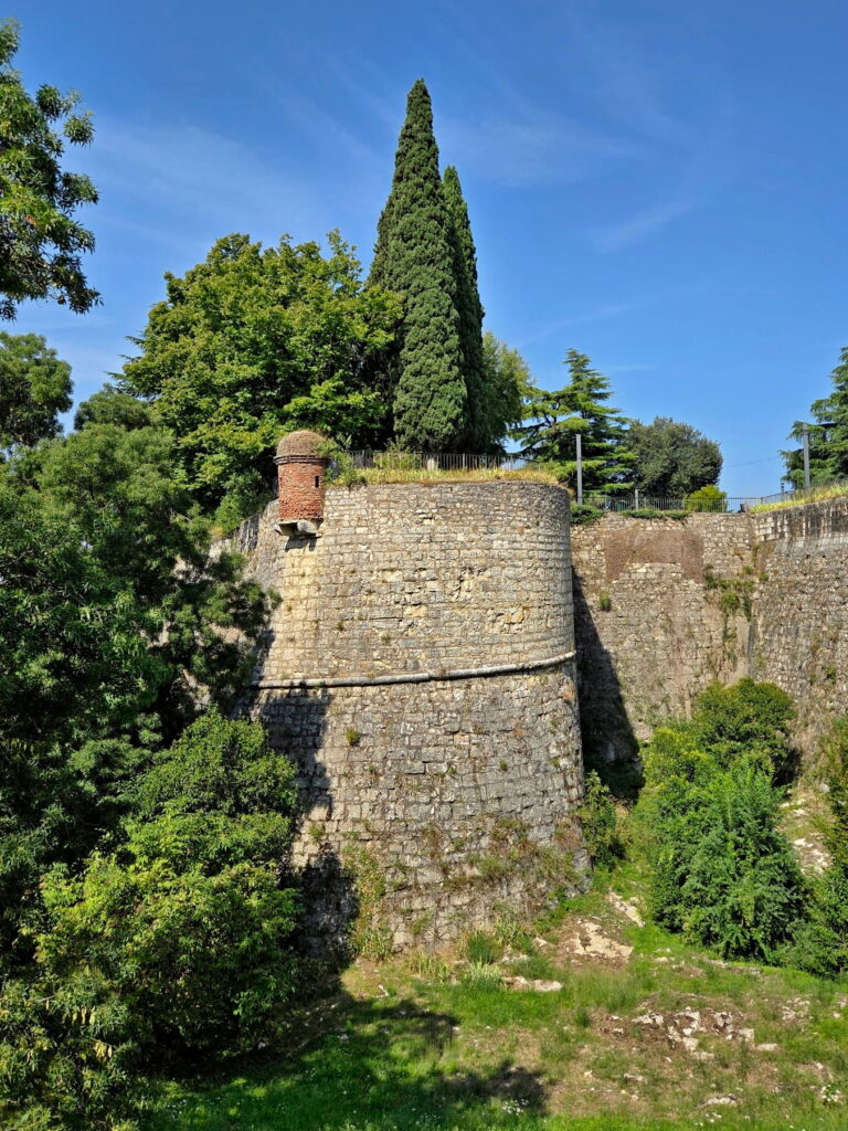 A rounded stone bastion of Brescia Castle with a small brick watchtower and lush green trees.