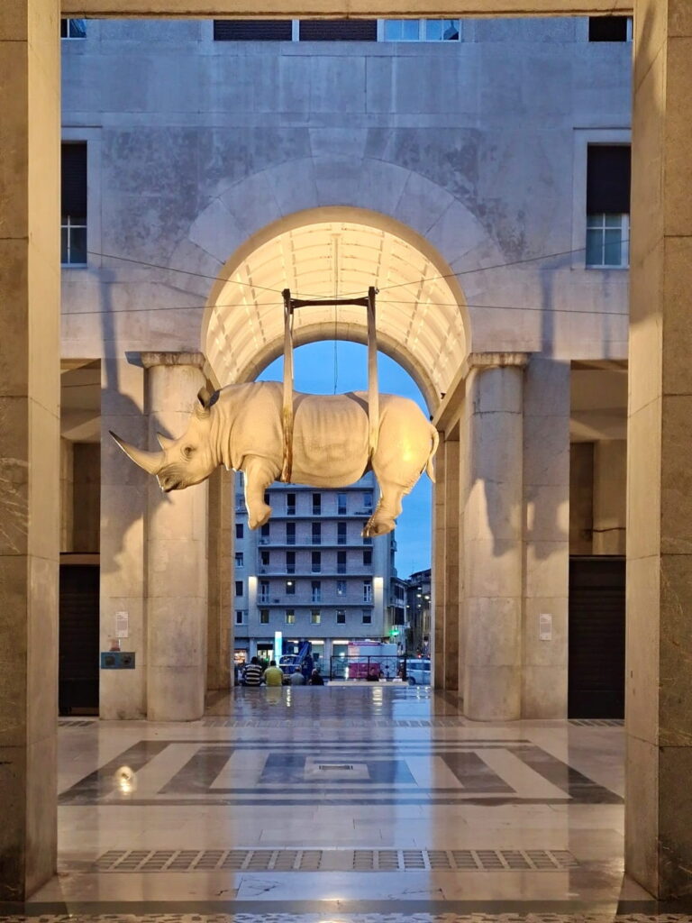 Hanging rhinoceros sculpture in Brescia, Italy, illuminated at dusk under an archway