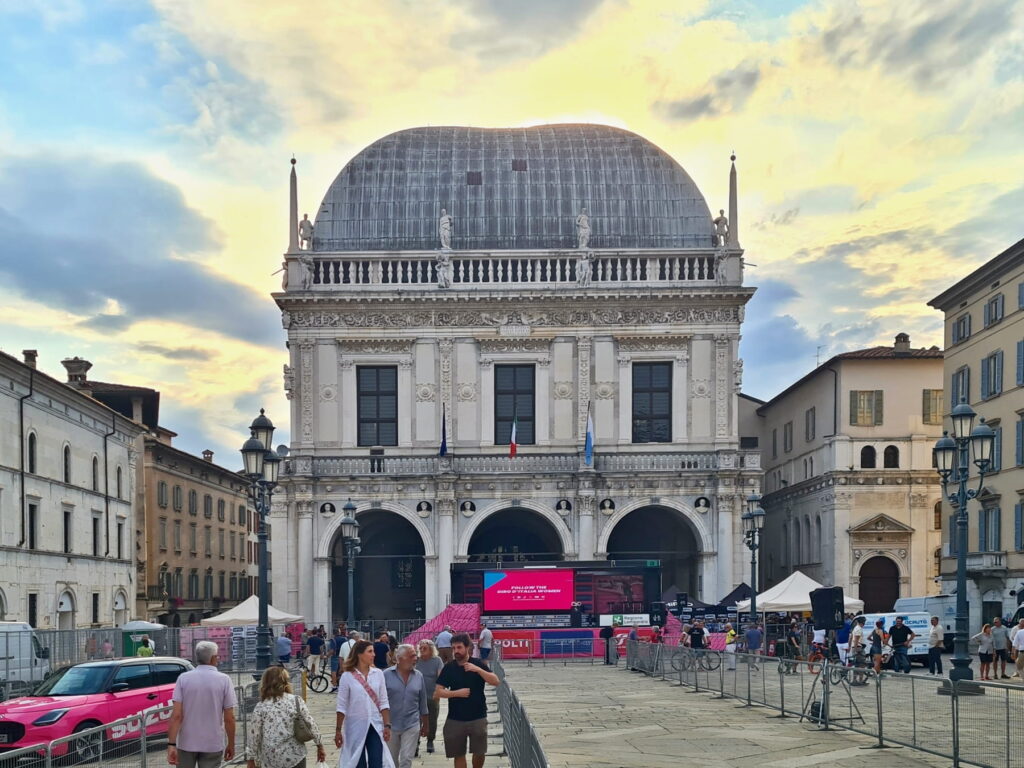 Palazzo della Loggia in Brescia at sunset with a large event screen and crowds in the square.