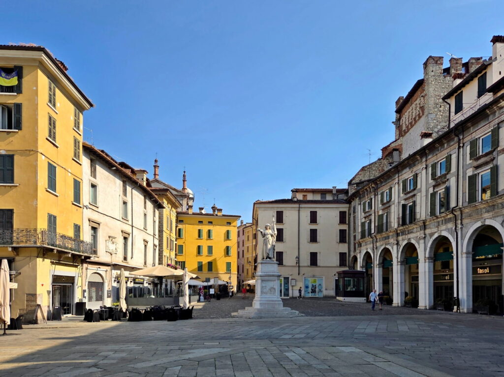 Wide view of Piazza della Loggia in Brescia with historic yellow buildings and a central monument.