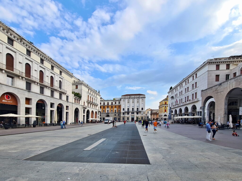 A wide perspective of the paved Piazza della Vittoria under a blue sky with scattered clouds in Brescia.