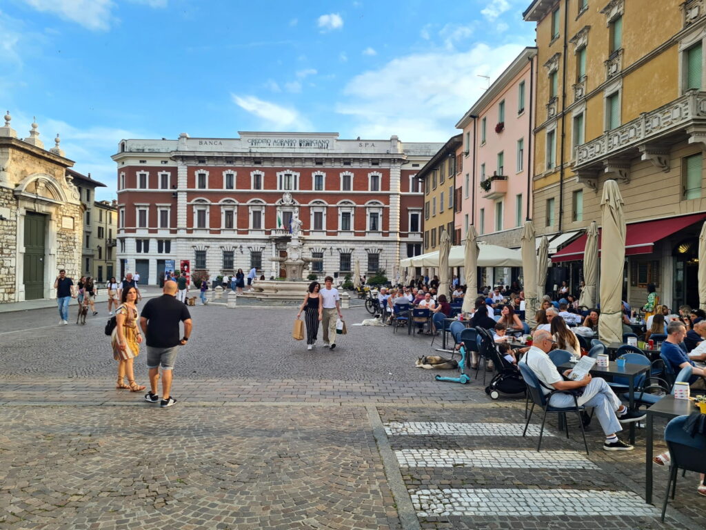 People sitting at outdoor cafe tables in Piazza Paolo VI with a grand historic bank building in the background.