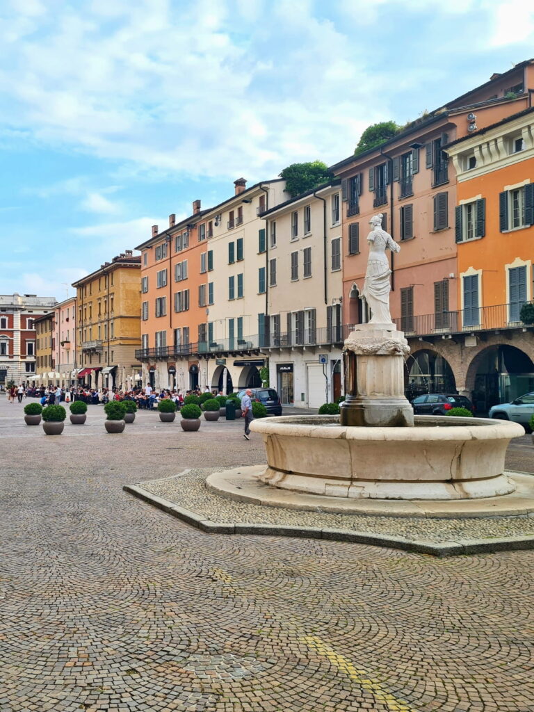A stone fountain with a marble statue in a paved square lined with colorful Italian apartment buildings.