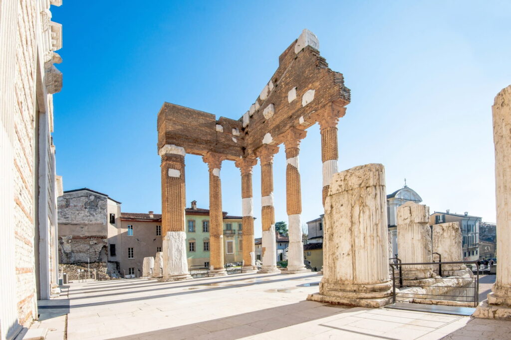 Standing Corinthian columns of the Roman Capitolium temple against a bright blue sky in Brescia, Italy.