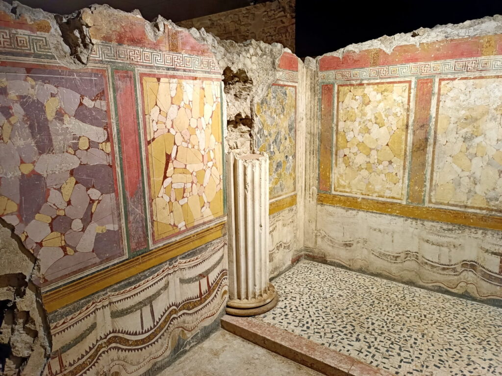 Interior of an ancient Roman room featuring vibrant geometric wall frescoes and a black and white mosaic floor.