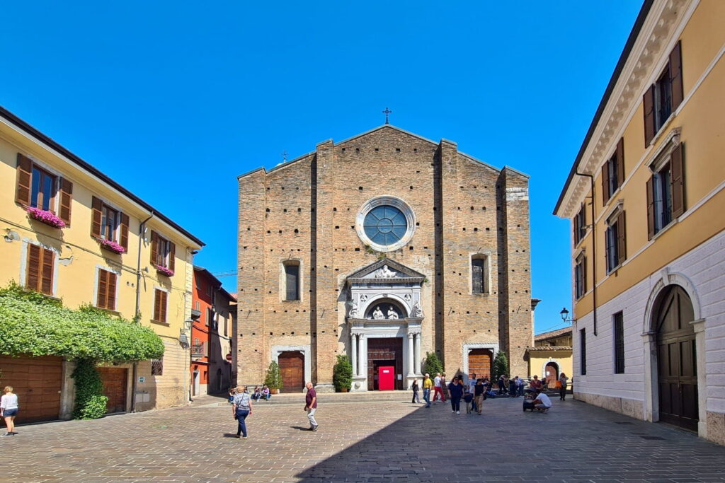 Wide view of the Duomo square in Salò, Italy, with colorful historic buildings and people walking in the sun.