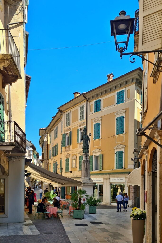 A tall statue on a pillar in a Salò square surrounded by yellow buildings and outdoor cafe seating.