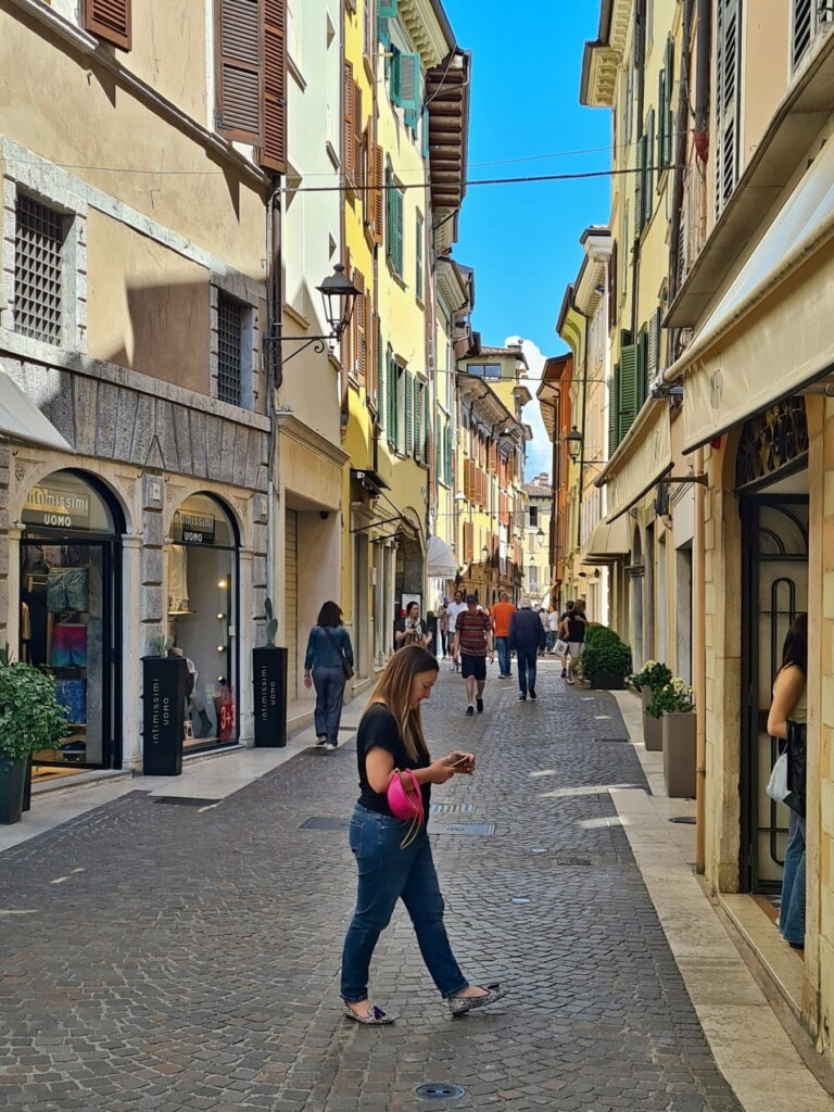 People walking along a narrow shopping street in Salò, Italy, featuring stone paving and boutique storefronts.