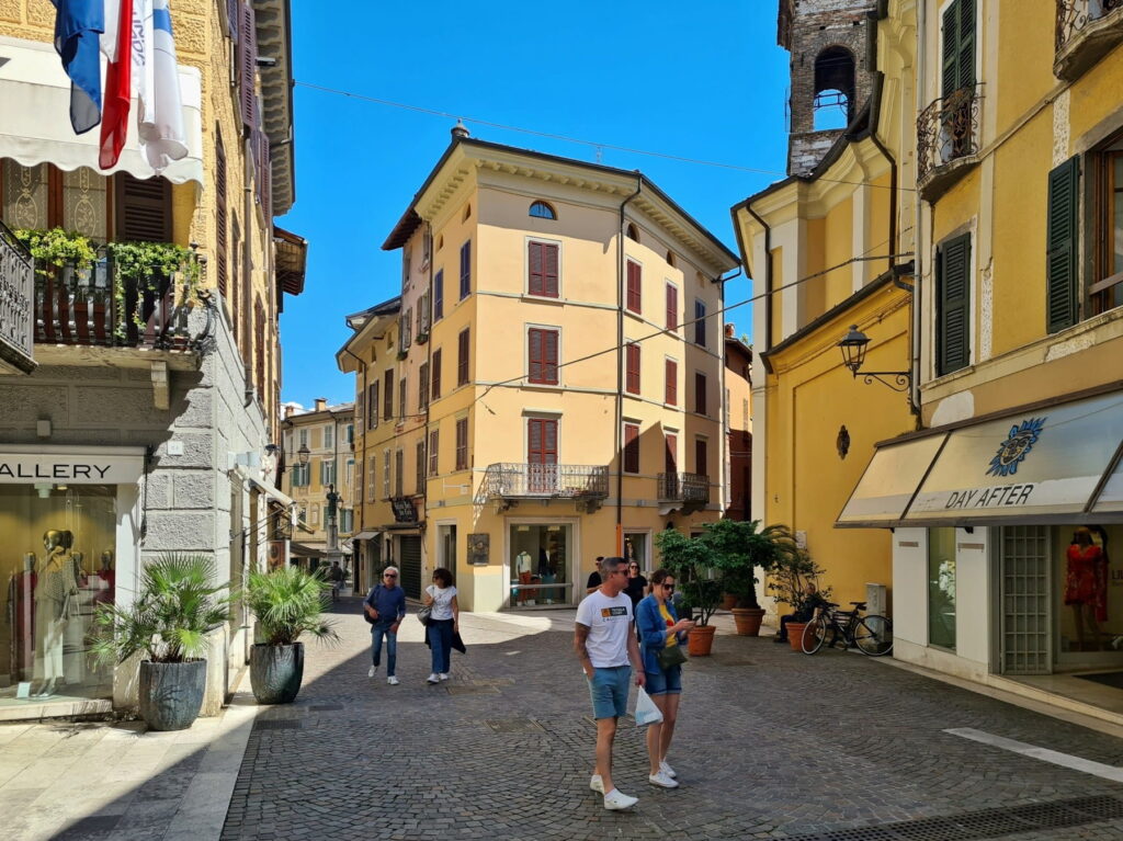 Travelers walking through a bright intersection in Salò's historic center with a church tower in the background.