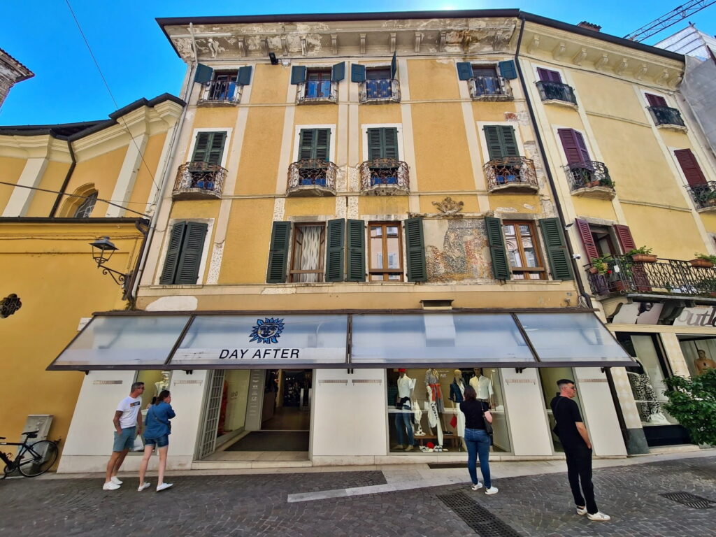 A yellow building in Salò with green shutters, a clothing store on the ground floor, and a faded wall fresco.