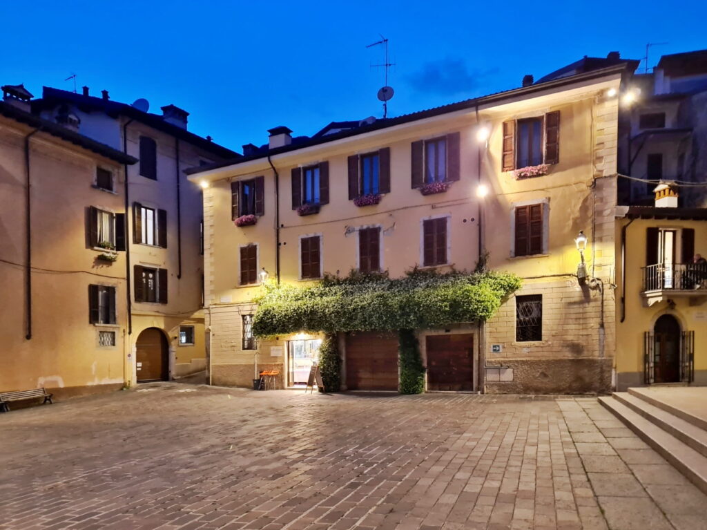 A quiet paved square in Salò at twilight with glowing streetlights and buildings draped in greenery.