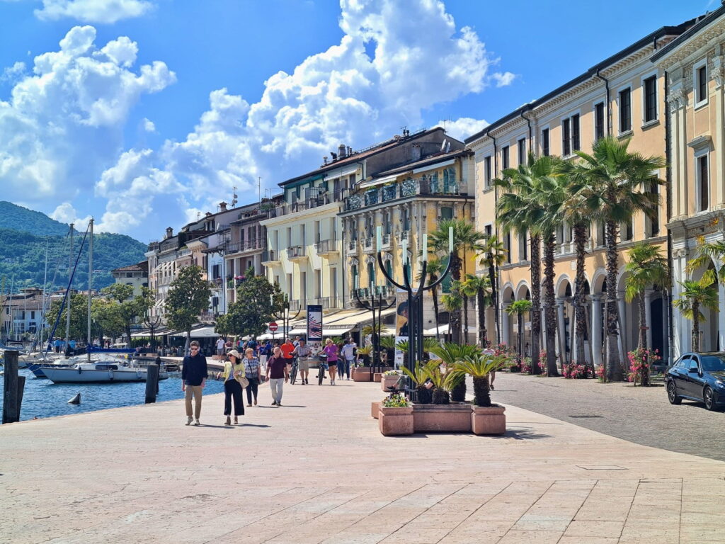 People walking along the sunny Lungolago Zanardelli in Salò, Italy, with palm trees and elegant historic buildings.
