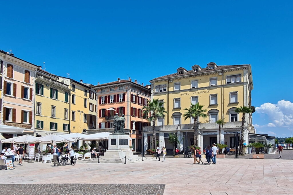 A spacious paved square along the Salò waterfront with a monument and the yellow facade of Hotel Metropole.