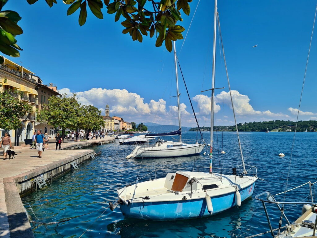 White sailboats moored in the blue water of Lake Garda next to the shaded promenade of Salò, Italy.