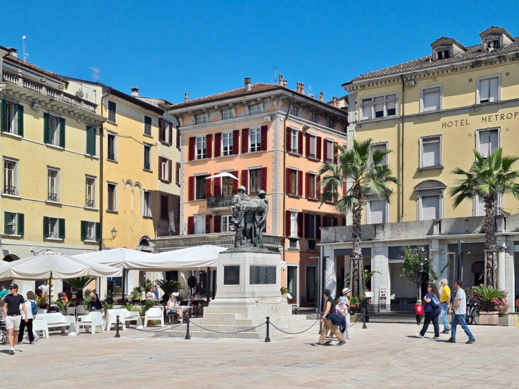 Close-up of the War Memorial in Piazza Vittoria, Salò, with historic buildings and outdoor cafe seating.