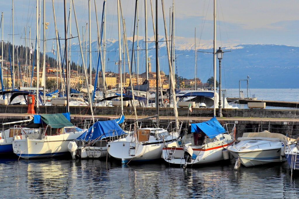Sailboats moored in Salò harbor with a view of snow-capped mountains across Lake Garda in Italy.