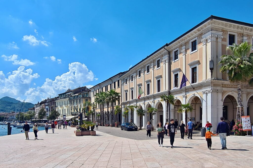 The grand Palazzo della Magnifica Patria with arched porticos and palm trees on the Salò waterfront in Italy.
