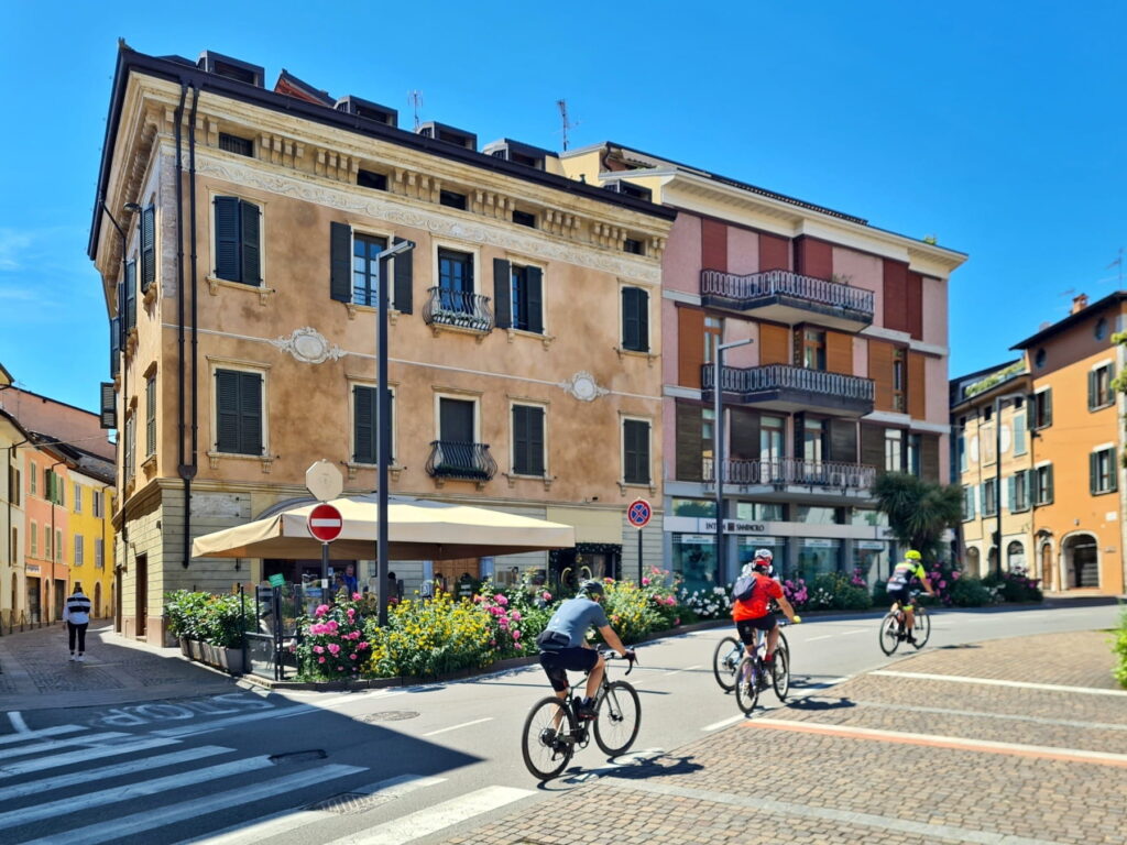 Cyclists riding past a historic building with flower beds in Salò, Italy, under a clear blue sky.