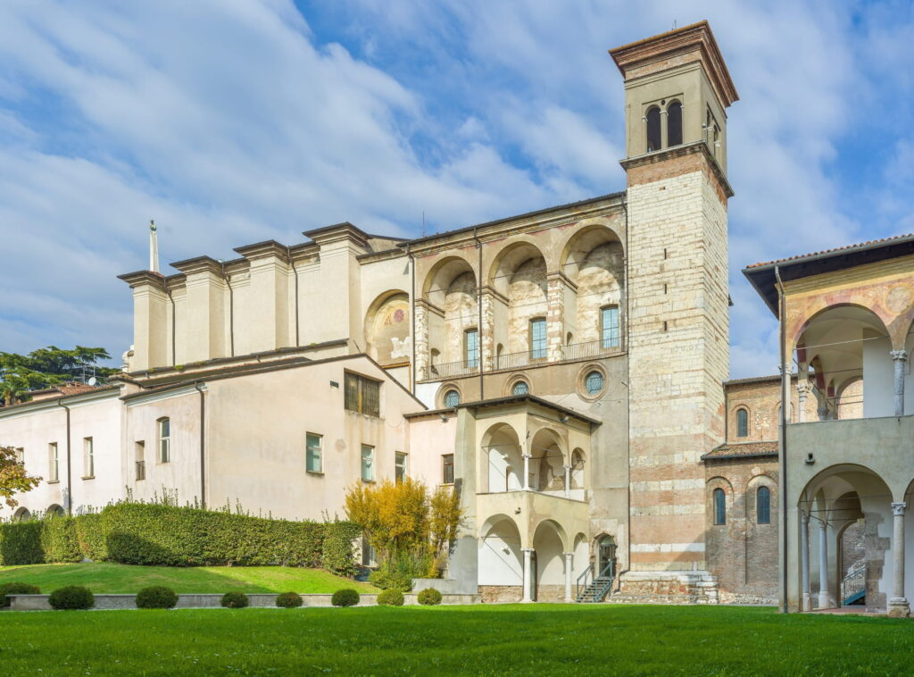 A green lawn and garden area with a view of the Romanesque church tower and stone arches of Santa Giulia.